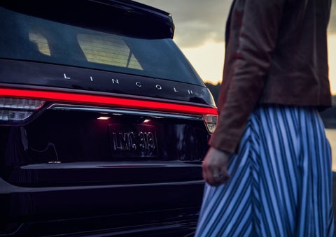 A person is shown near the rear of a 2024 Lincoln Aviator® SUV as the Lincoln Embrace illuminates the rear lights | Libertyville Lincoln Sales, Inc. in Libertyville IL